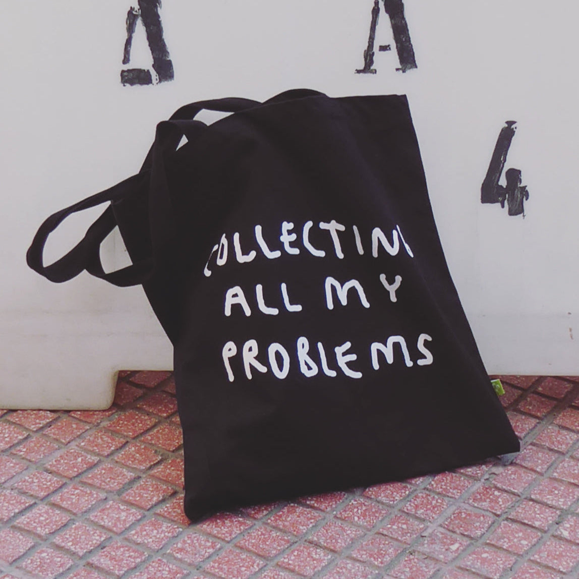 Schwarze Jutetasche mit weißem Motiv Collecting all my Problems. Die Tasche lehnt an einer weißen Wand.