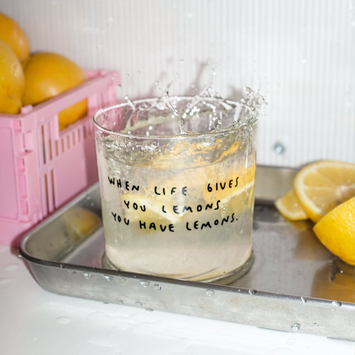 Wasserglas mit Schriftzug when life gives you lemons. Das Glas ist mit Wsser befüllt und eine Zitrone ist in das Glas gefallen. Im Hintergrund sind Zitronen abgelegt.