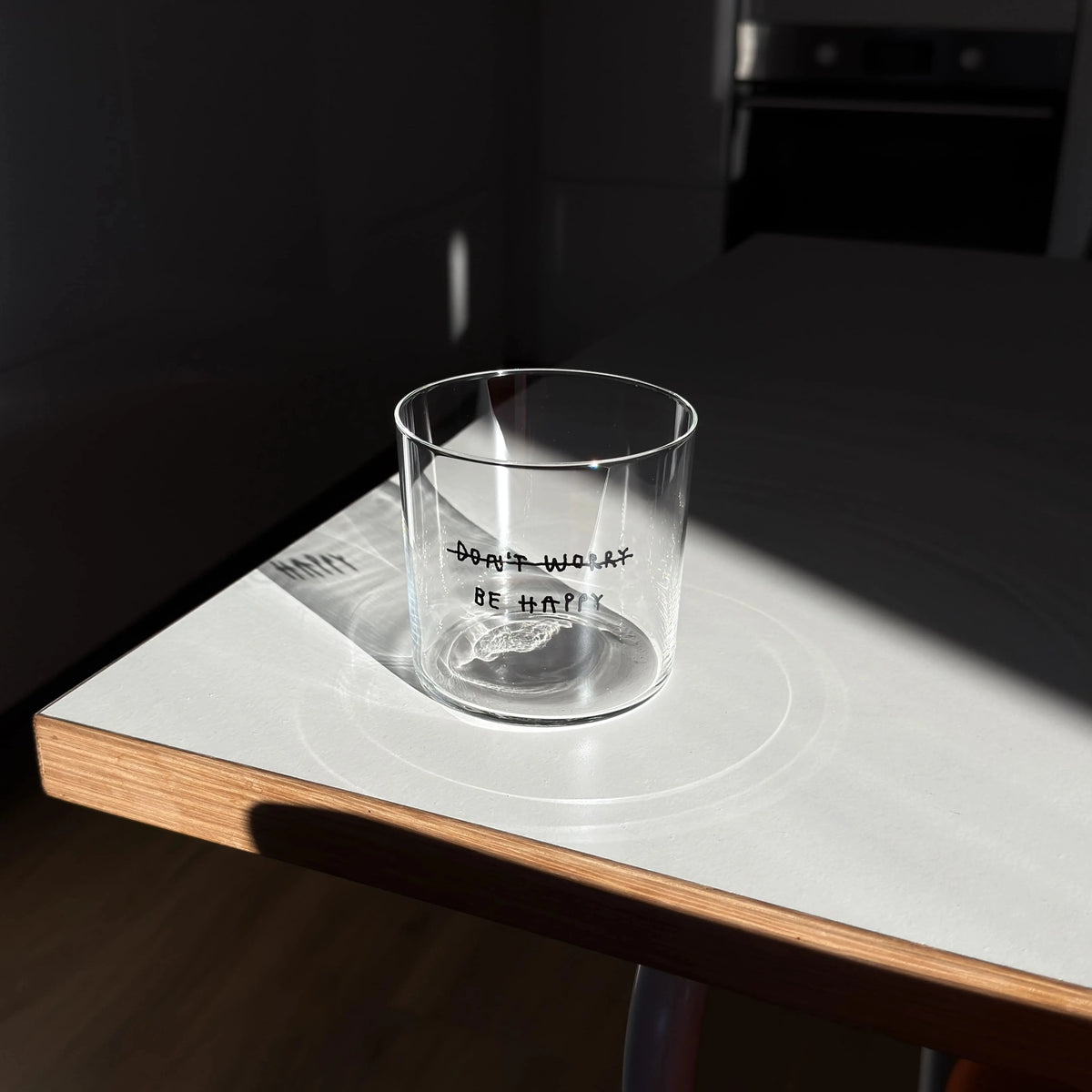 Transparentes Medium Designer Glas mit schwarzem Schriftzug "DONT WORRY BE HAPPY" wobei DONT WORRY durchgestrichen ist und lediglich die Aufforderung BE HAPPY im Vordergrund steht, die Gläser haben dünnen Wänden und Boden. 
 
Das Glas ist unbefüllt auf einer weißen Tischpaltte. 