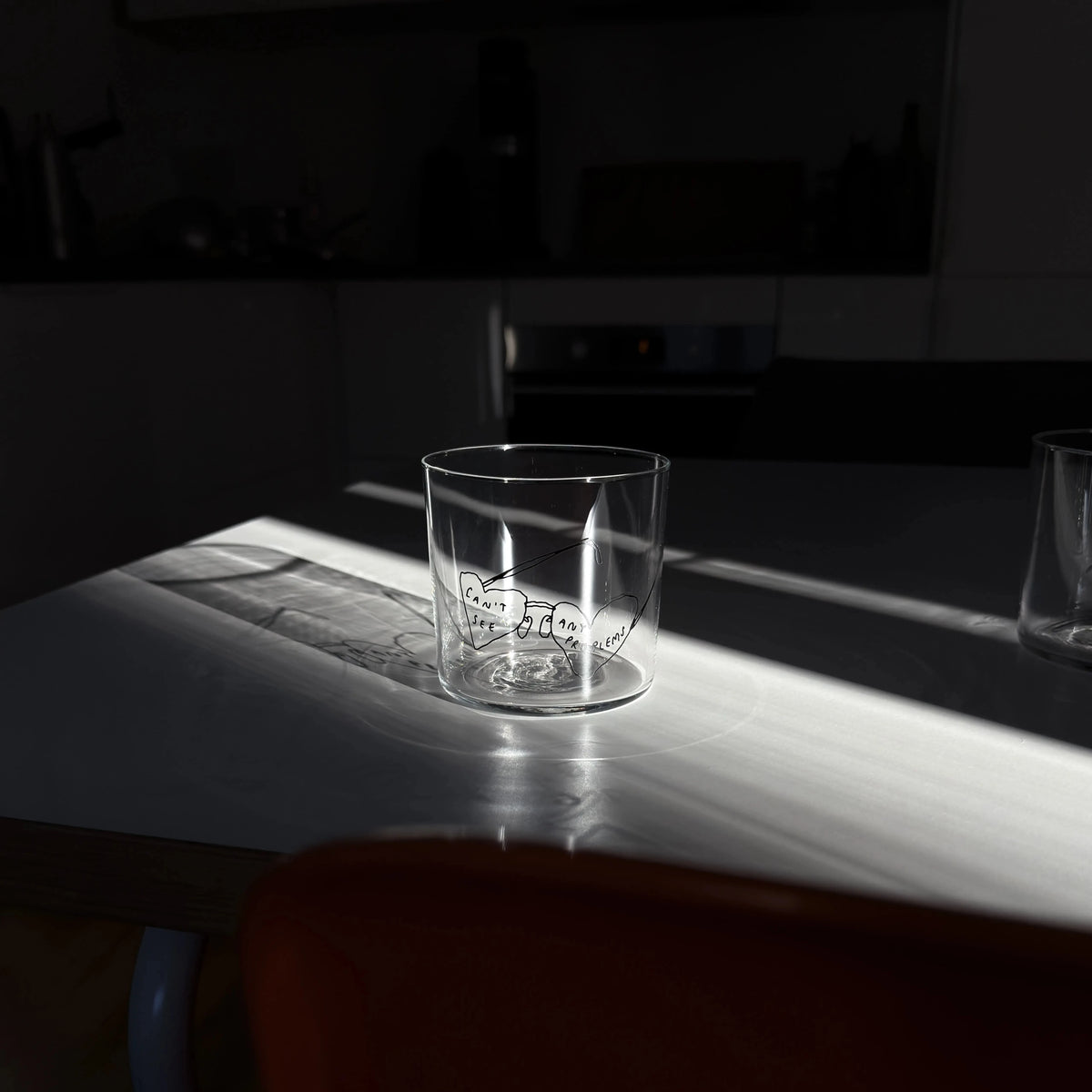 Transparentes Medium Designer Glas mit schwarzem Schriftzug "can't see any problems" mit einer Brille, die Brillengläser in herzformen hat. Das Glas steht in einem Lichtkegel, viel Schatten im Hintergrund, auf einem weißen Tisch.