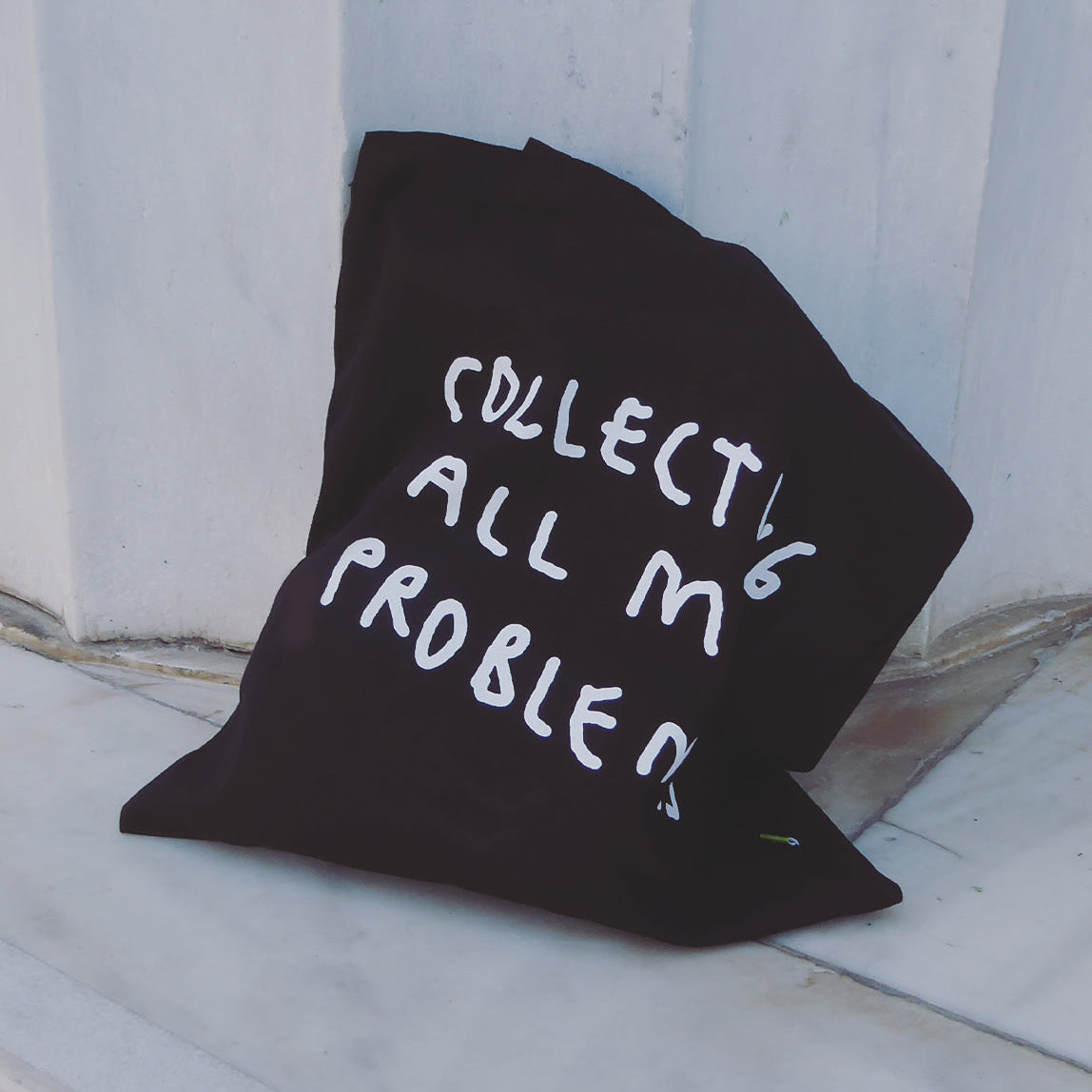 Schwarze Jutetasche mit weißem Motiv Collecting all my Problems. Die Tasche lehnt an einer Mauer und liegt auf dem Boden.