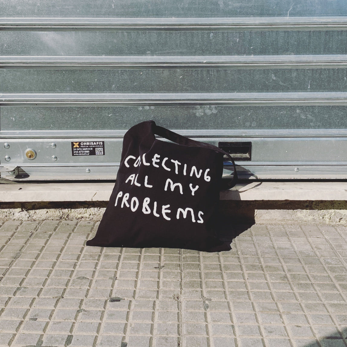 Schwarze Jutetasche mit weißem Motiv Collecting all my Problems. Die Tasche liegt auf der Straße vor einem Garagentor.