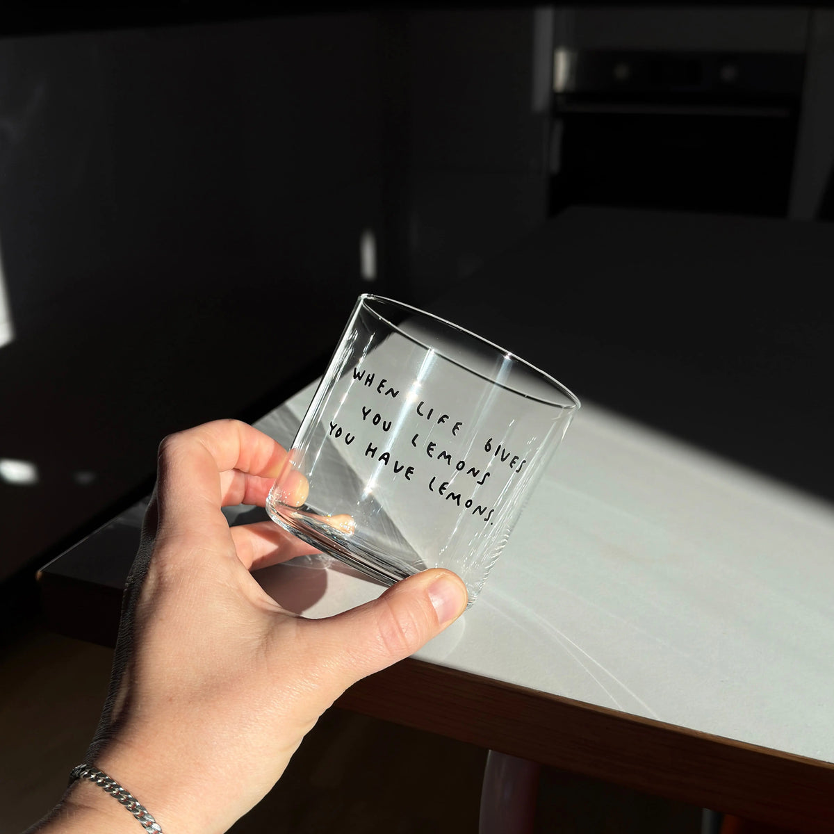 Transparentes Medium Designer Glas mit schwarzem Schriftzug "when life gives you lemons" mit dünnen Wänden und Boden. Das Glas steht im Licht auf einem weißen Tisch und wirft einen weiten Schatten. Das Glas ist mit Milch befüllt. 