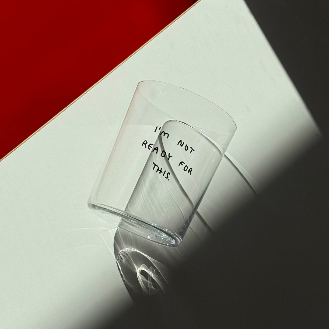 Großes Designer-Trinkglas mit sehr dünnen Glaswänden und einer schwarzen, einfarbigen Illustration auf der Vorderseite. Das Motiv zeigt ein Glas unter Wasser mit einem Schwamm darin und den Text „I’m not ready for this“. Illustration von Johanna Schwarzer.

Das Glas liegt auf einem weißen Tisch mit einem Orangenen Stuhl daneben.