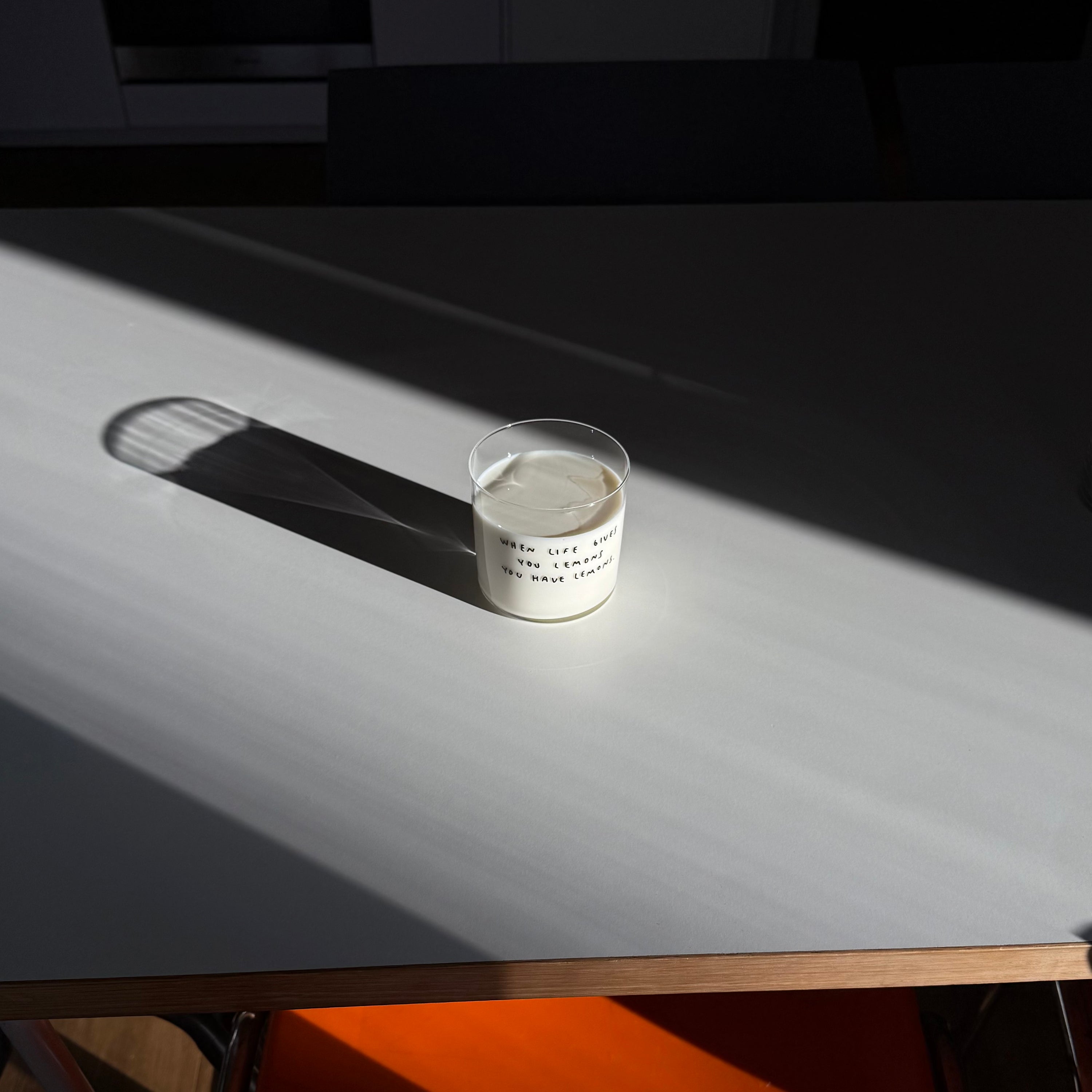 Transparentes Medium Designer Glas mit schwarzem Schriftzug "when life gives you lemons" mit dünnen Wänden und Boden. Das Glas steht im Licht auf einem weißen Tisch und wirft einen weiten Schatten. Das Glas ist mit Milch befüllt. 