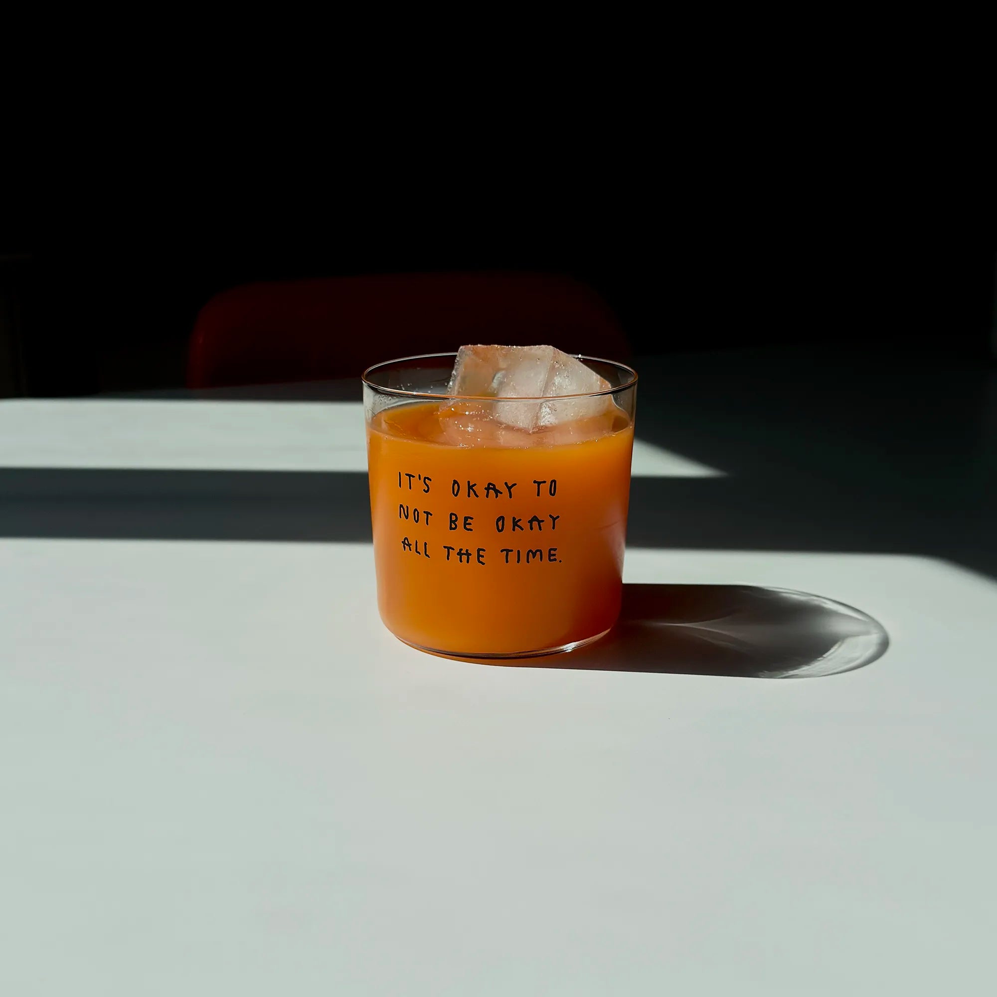 Transparentes Medium Designer Glas mit schwarzem Schriftzug "It's okay to not be okay all the time" mit dünnen Wänden und Boden. 

Das Glas ist mit einer orangenen Flüssigkeit befüllt und Eiswürfeln. Es steht auf einem weißen Tisch und wirft einen schatten.