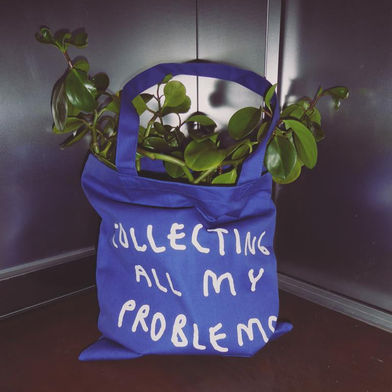 Blaue Jutetasche mit weißem Motiv "Collecting all my problems". Die Tasche steht in einem Aufzug und ist mit einer Pflanze befüllt.