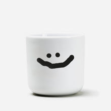 Weißer henkelloser Porzellan-Becher mit einer einfachen schwarzen Illustration eines großen grinsenden Smiley.