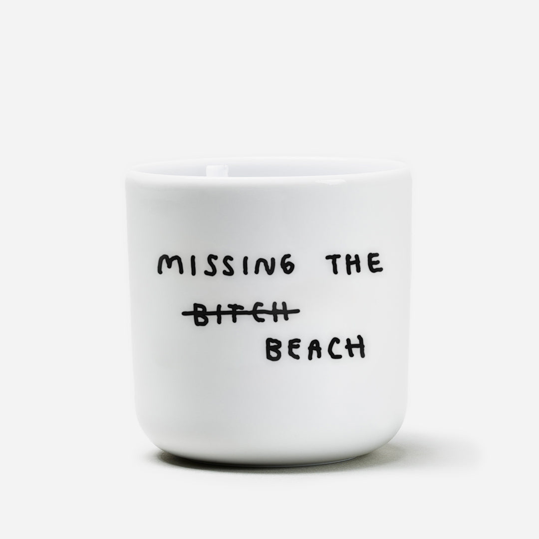 Weißer Becher aus Porzellan, Mittelgroß mit schwarzem Spruch "Missing the beach" auf Weißen Hintergrund.