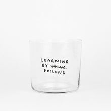 Klares, zylindrisches Trinkglas mit schwarzem, handschriftlich wirkendem Text. Darauf steht „Learning by doing“, wobei das Wort „doing“ durchgestrichen ist und darunter „failing“ steht, sodass der Satz humorvoll zu „Learning by failing“ geändert wird.