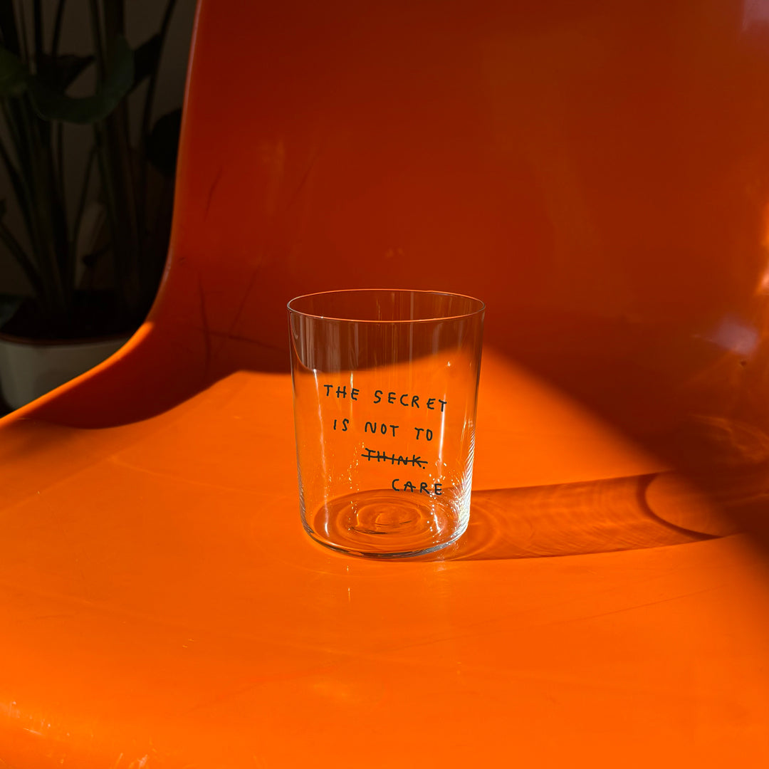 Großes Designer-Trinkglas mit sehr dünnen Glaswänden und einer schwarzen, einfarbigen Illustration auf der Vorderseite. Das Motiv auf dem Glas ist eine Handschrift in Schwarz in der geschrieben ist The secret is not to think wobei das think durchgestrichen ist und drunter das Wort CARE steht. Das Glas steht auf einem orangenen Stuhl.