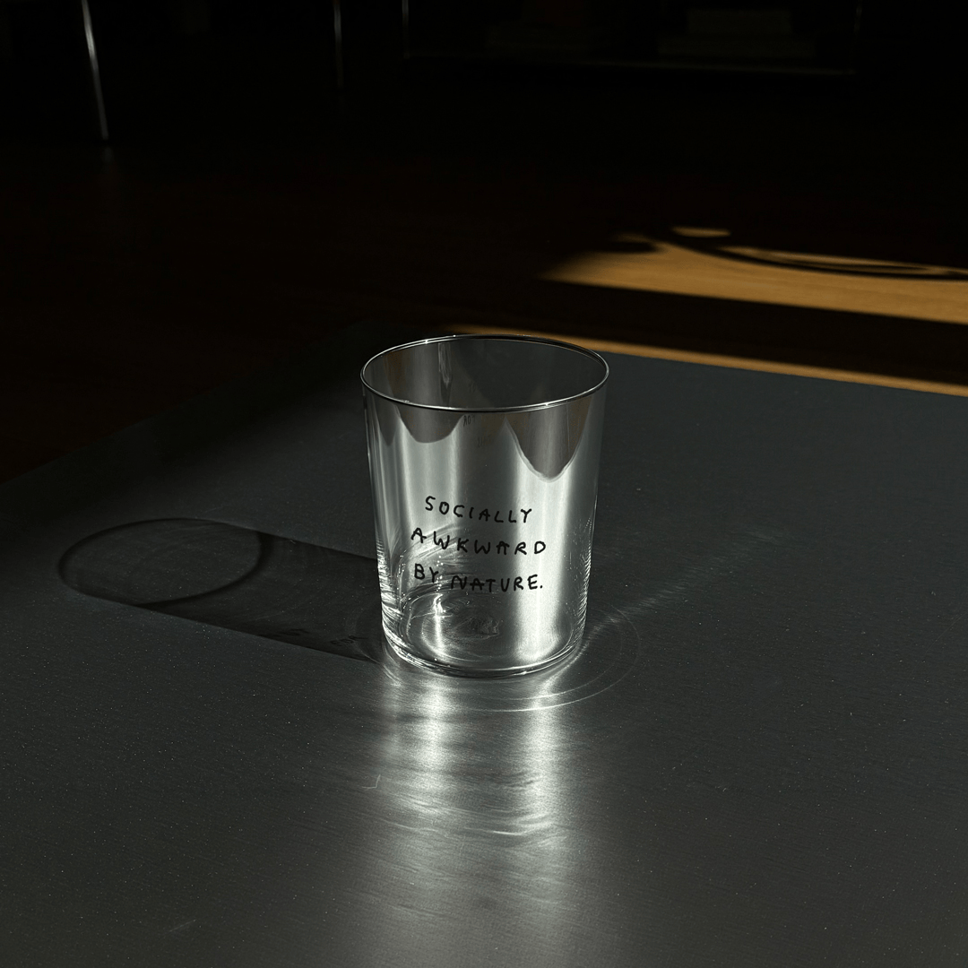Großes Designer-Trinkglas mit sehr dünnen Glaswänden und einer schwarzen, einfarbigen Illustration auf der Vorderseite. Das Motiv auf dem Glas ist eine Handschrift in Schwarz in der geschrieben ist SOCIALLY AWKWARD BY NATURE. Das Glas steht auf einer Metallplatte.