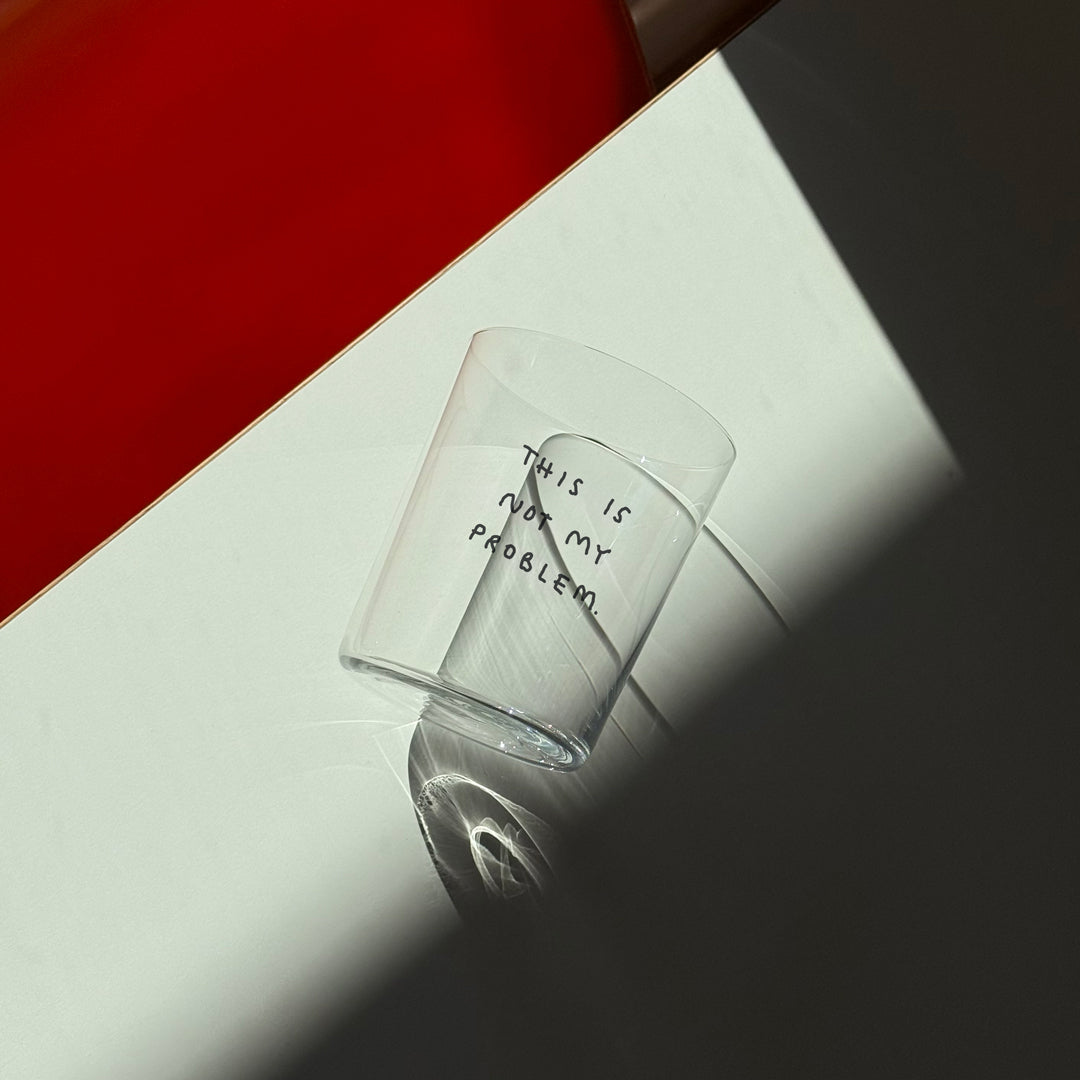 Großes Designer-Trinkglas mit sehr dünnen Glaswänden und einer schwarzen, einfarbigen Illustration auf der Vorderseite. Das Motiv auf dem Glas ist eine Handschrift in Schwarz in der geschrieben ist THIS IS NOT MY PROBLEM, auf weißem Tisch neben dem ein orangener Stuhl steht.