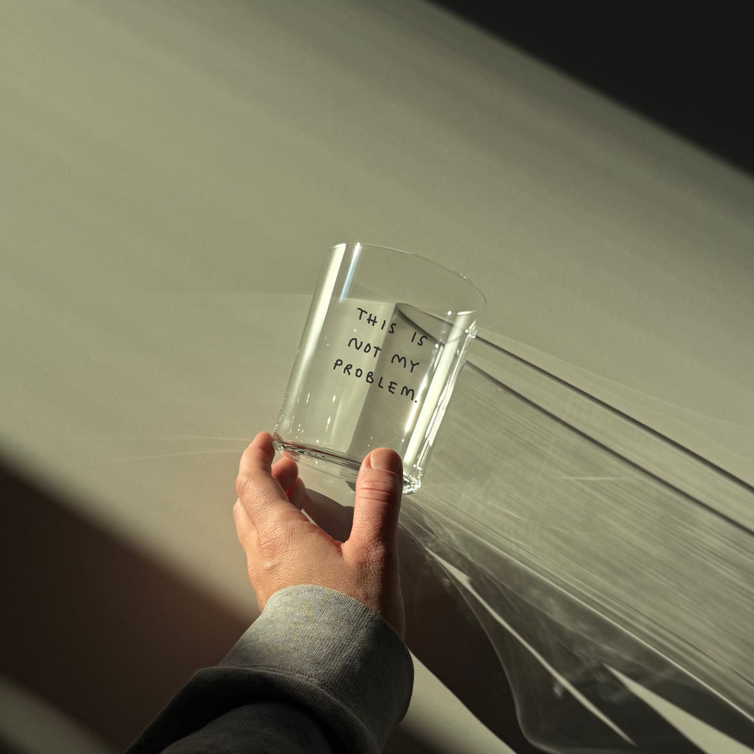 Großes Designer-Trinkglas mit sehr dünnen Glaswänden und einer schwarzen, einfarbigen Illustration auf der Vorderseite. Das Motiv auf dem Glas ist eine Handschrift in Schwarz in der geschrieben ist THIS IS NOT MY PROBLEM, auf weißem Tisch und von einer Hand gehalten.