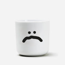 Weißer henkelloser Porzellan-Becher mit einer einfachen schwarzen Illustration eines großen traurigen Smiley auf weißen Hintergrund.