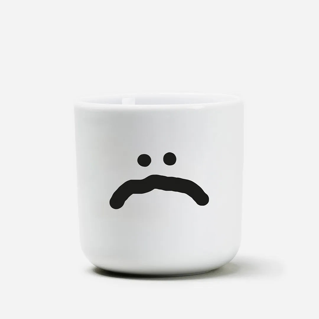 Weißer henkelloser Porzellan-Becher mit einer einfachen schwarzen Illustration eines großen traurigen Smiley auf weißen Hintergrund.