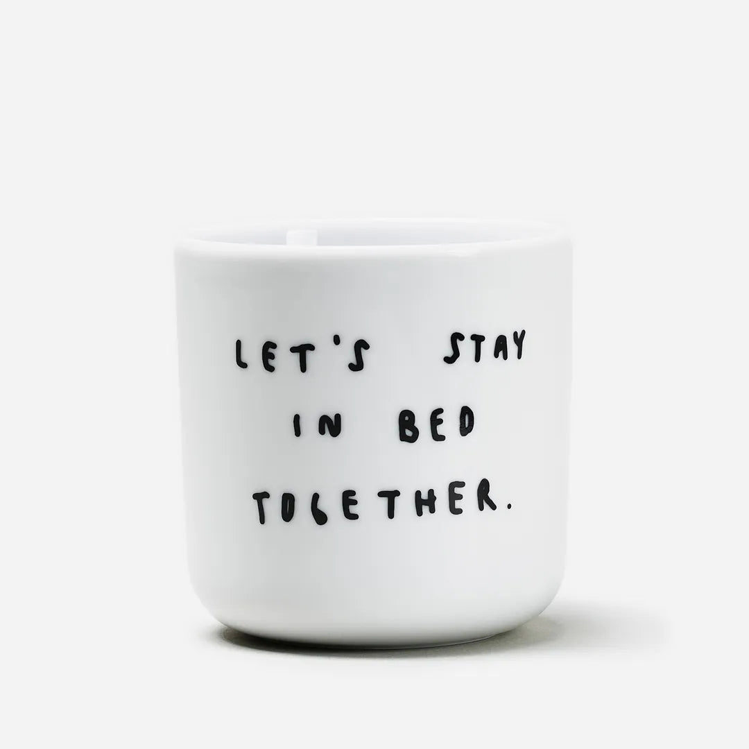 Weißer henkelloser Porzellan-Becher mit einer einfachen schwarzen Illustration, einem handschriftlichen Text der besagt "LET'S STAY IN BED TOGETHER" auf weißen Hintergrund.