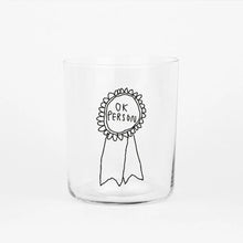 Großes Designer-Trinkglas mit sehr dünnen Glaswänden und einer schwarzen, einfarbigen Illustration auf der Vorderseite. Das Motiv zeigt eine Brosche inklusive einer Typografie in der geschrieben ist OK PERSON auf weißem Hintergrund.