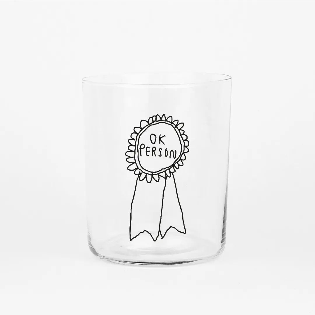 Großes Designer-Trinkglas mit sehr dünnen Glaswänden und einer schwarzen, einfarbigen Illustration auf der Vorderseite. Das Motiv zeigt eine Brosche inklusive einer Typografie in der geschrieben ist OK PERSON auf weißem Hintergrund.