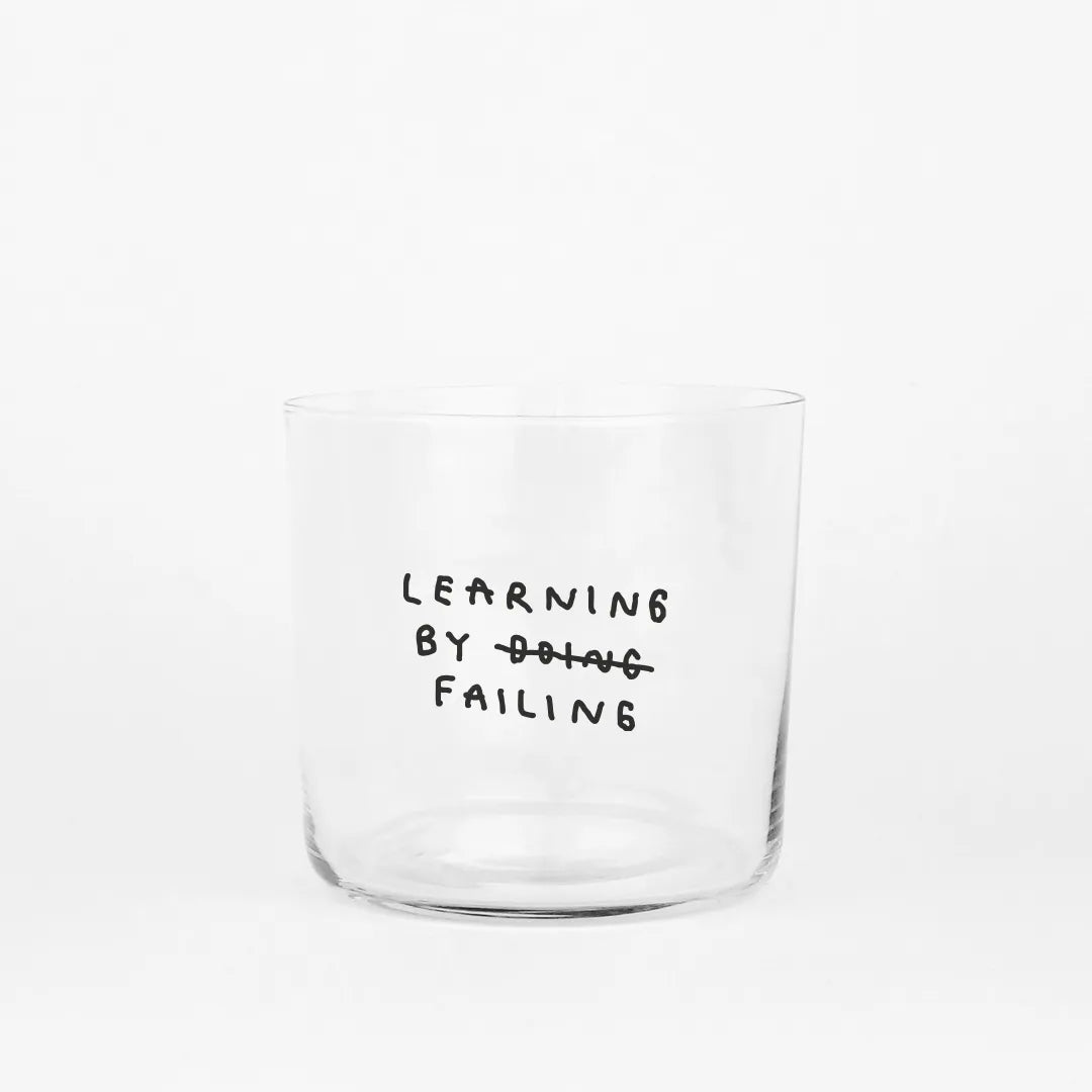 Klares, zylindrisches Trinkglas mit schwarzem, handschriftlich wirkendem Text. Darauf steht „Learning by doing“, wobei das Wort „doing“ durchgestrichen ist und darunter „failing“ steht, sodass der Satz humorvoll zu „Learning by failing“ geändert wird.
