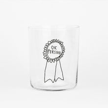 Großes Designer-Trinkglas mit sehr dünnen Glaswänden und einer schwarzen, einfarbigen Illustration auf der Vorderseite. Das Motiv zeigt eine Brosche inklusive einer Typografie in der geschrieben ist OK PERSON auf weißem Hintergrund.