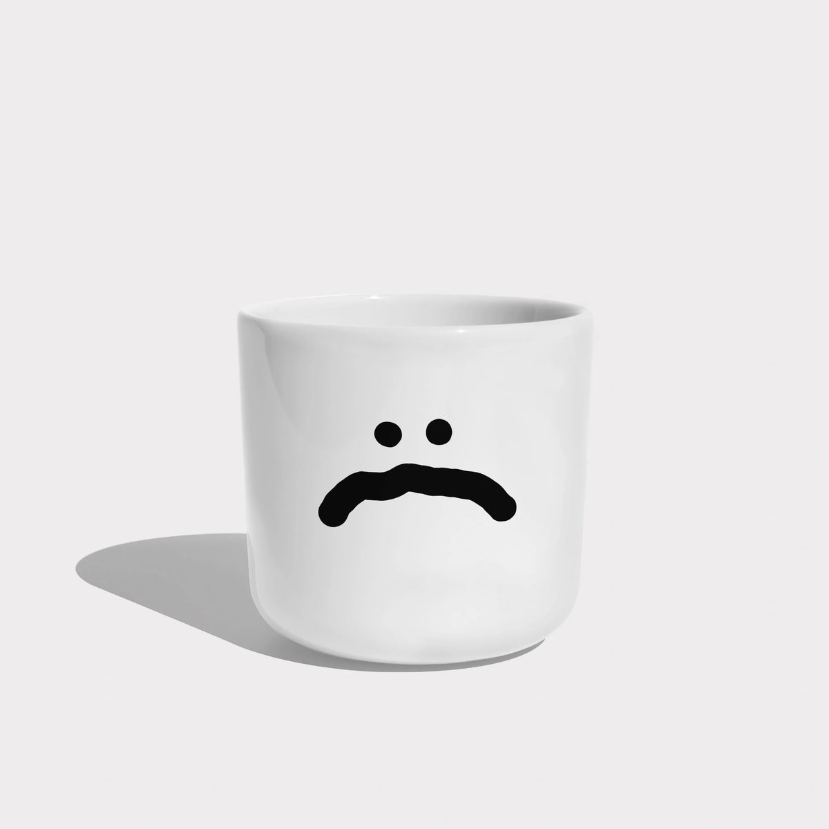 Weißer henkelloser Porzellan-Becher mit einer einfachen schwarzen Illustration eines großen traurigen Smiley auf weißen Hintergrund.
