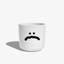 Weißer henkelloser Porzellan-Becher mit einer einfachen schwarzen Illustration eines großen traurigen Smiley auf weißen Hintergrund.