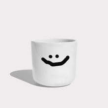 Weißer henkelloser Porzellan-Becher mit einer einfachen schwarzen Illustration eines großen grinsenden Smiley.