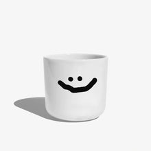 Weißer henkelloser Porzellan-Becher mit einer einfachen schwarzen Illustration eines großen grinsenden Smiley.