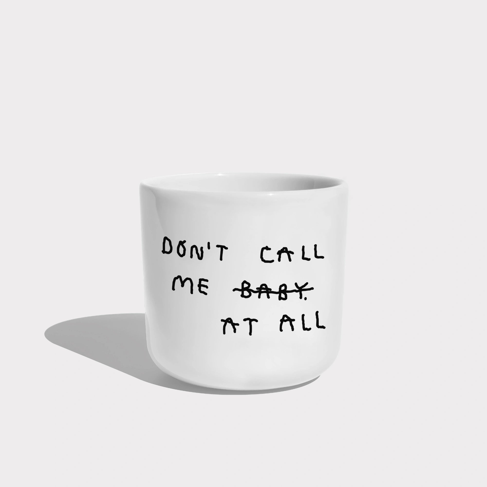 Weißer henkelloser Porzellan-Becher mit schwarzen Text „don't call me at baby" - das Baby ist durchgestrichen und durch "at all" ersetzt. Zu Deutsch (Nenn mich nicht Baby, bzw. Ruf mich nicht an).

Der Becher ist auf weißem Hintergrund.