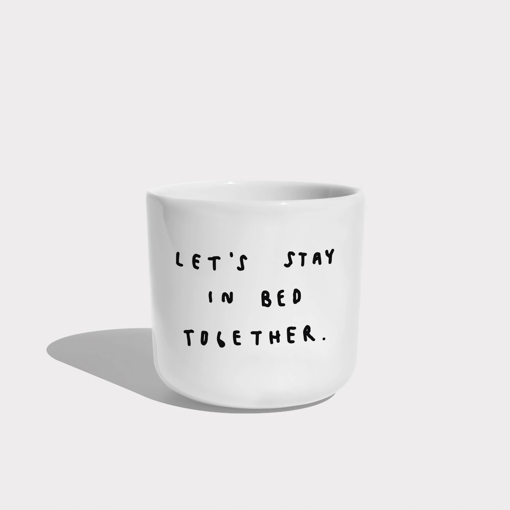 Weißer henkelloser Porzellan-Becher mit einer einfachen schwarzen Illustration, einem handschriftlichen Text der besagt "LET'S STAY IN BED TOGETHER" auf weißen Hintergrund.