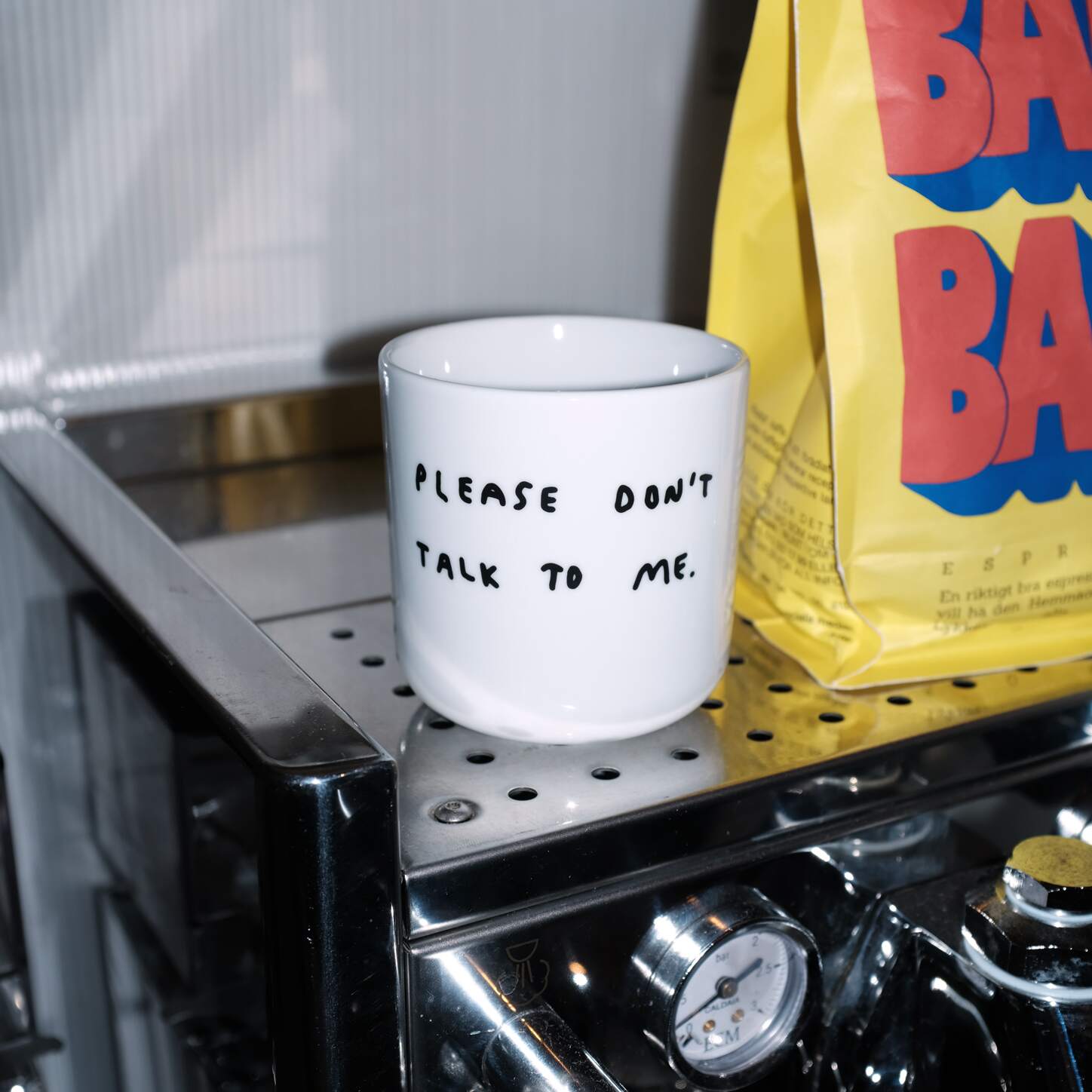 Weißer henkelloser Porzellan-Becher mit einer einfachen schwarzen Illustration, einem handschriftlichen Text der besagt "PLEASE DON'T TALK TO ME" auf weißen Hintergrund liegend. Der Becher steht auf einer Kaffeemaschine, daneben eine gelbe Packung Kaffee.