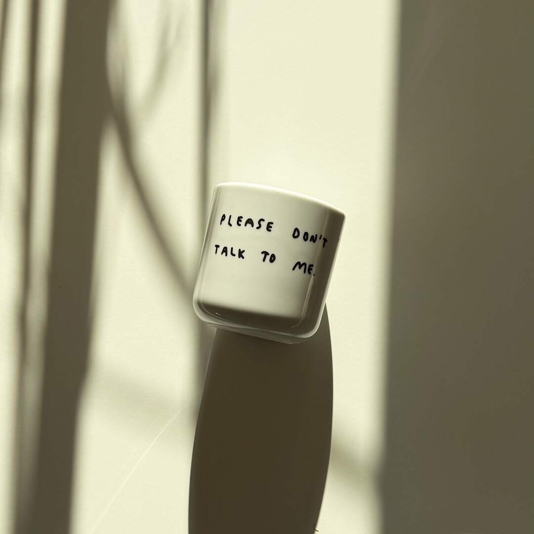 Weißer henkelloser Porzellan-Becher mit einer einfachen schwarzen Illustration, einem handschriftlichen Text der besagt "PLEASE DON'T TALK TO ME" auf weißen Hintergrund liegend. Der Becher liegt in einem milden Licht und wirft einen langen Schatten.