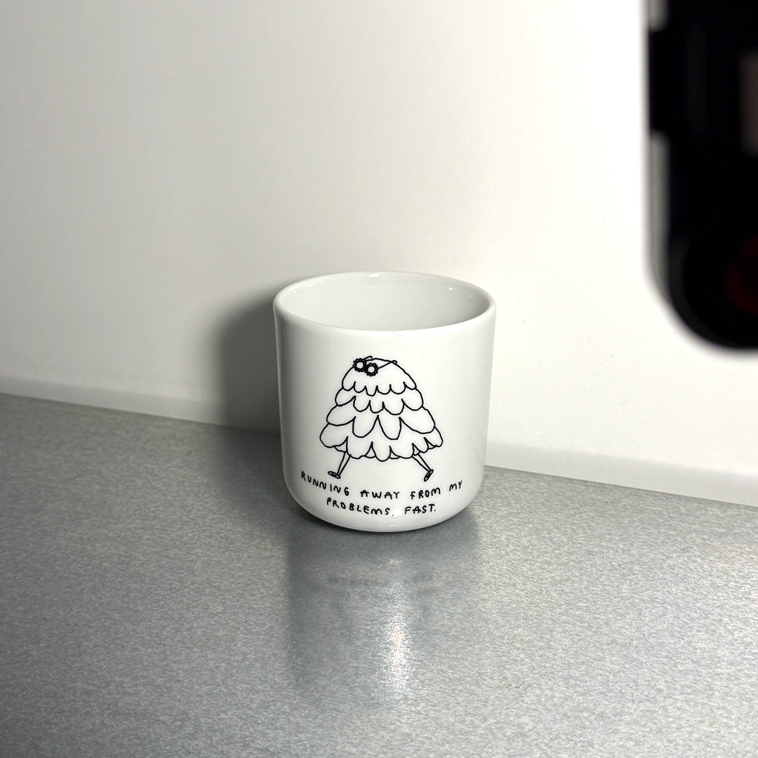 Weißer henkelloser Porzellan-Becher mit einer schwarzen Cartoon-Zeichnung einer flauschigen Figur und dem Text „Running away from my problems, fast“. Der Becher steht auf einem hellen Tisch im Sonnenlicht vor einer weißen Wand.