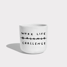 Weißer henkelloser Porzellan-Becher mit einer einfachen schwarzen Illustration, einem handschriftlichen Text der besagt "WORK LIFE CHALLENGE" wobei auch das Wort Balance dabeisteht und humorvoll durchgestrichen wird, dadurch von Challenge ersetzt wird. Auf weißen Hintergrund.