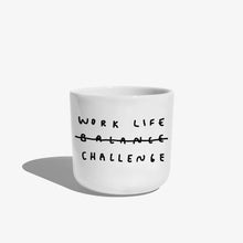 Weißer henkelloser Porzellan-Becher mit einer einfachen schwarzen Illustration, einem handschriftlichen Text der besagt "WORK LIFE CHALLENGE" wobei auch das Wort Balance dabeisteht und humorvoll durchgestrichen wird, dadurch von Challenge ersetzt wird. Auf weißen Hintergrund.