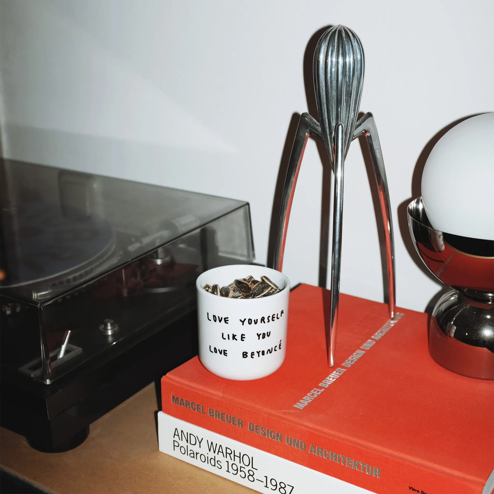 Weißer henkelloser Porzellan-Becher mit einer einfachen schwarzen Illustration, einem handschriftlichen Text der besagt "LOVE YOURSELF LIKE YOU LOVE BEYONCÉ". Die Tasse steht auf zwei Büchern von Andy Warhol und Marcel Breuer, daneben steht ein Schalplattenspieler und eine Lampe.