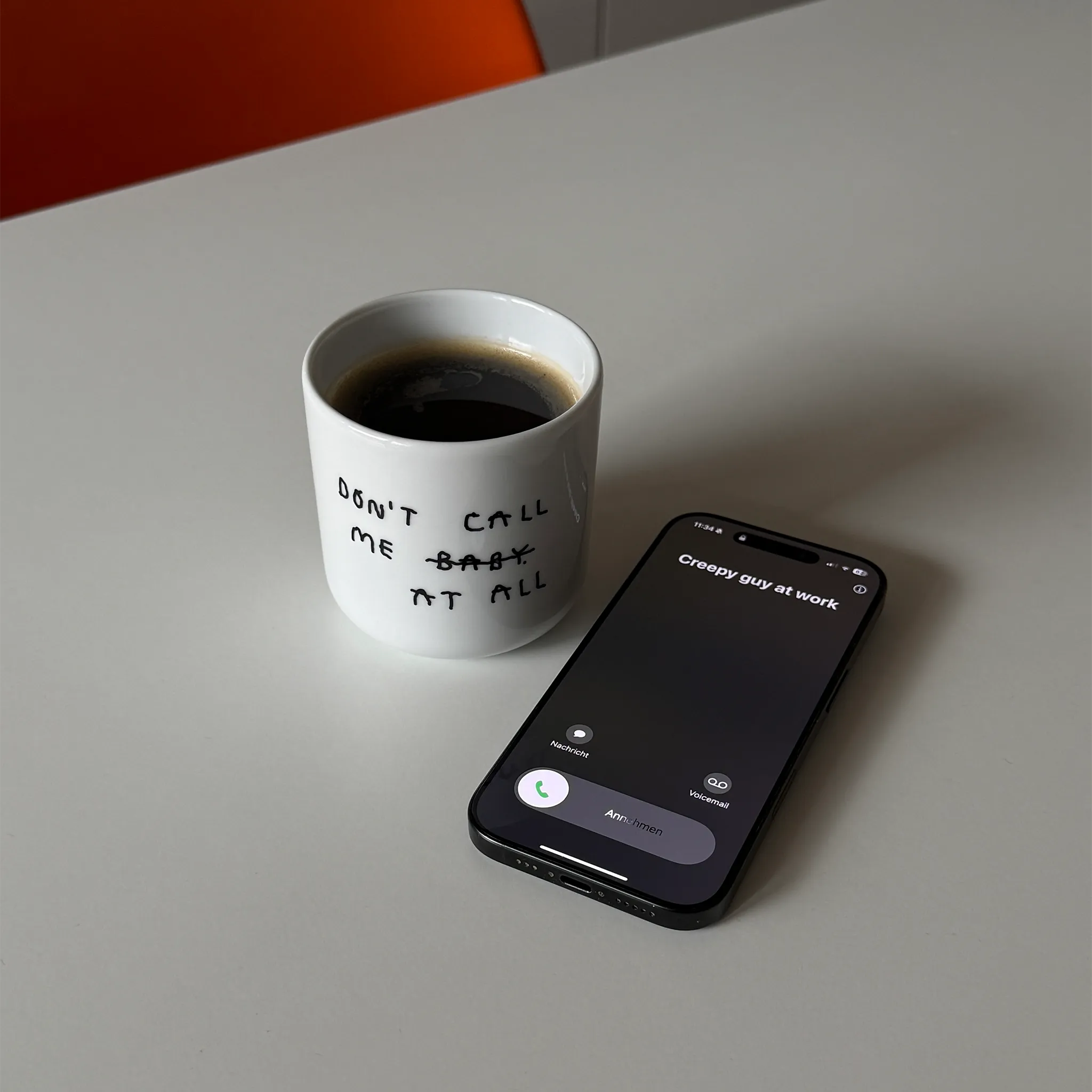 Weißer henkelloser Porzellan-Becher mit schwarzen Text „don't call me at baby" - das Baby ist durchgestrichen und durch "at all" ersetzt. Zu Deutsch (Nenn mich nicht Baby, bzw. Ruf mich nicht an). Der Becher steht auf einem Tisch in angenehmen Licht, ein orangener Stuhl ist im Hintergrund. Daneben liegt ein Iphone mit einem eingehenden Anruf von "creepy guy at work" im Bezug auf das Catcalling Motiv dem Becher, befüllt mit Kaffee.