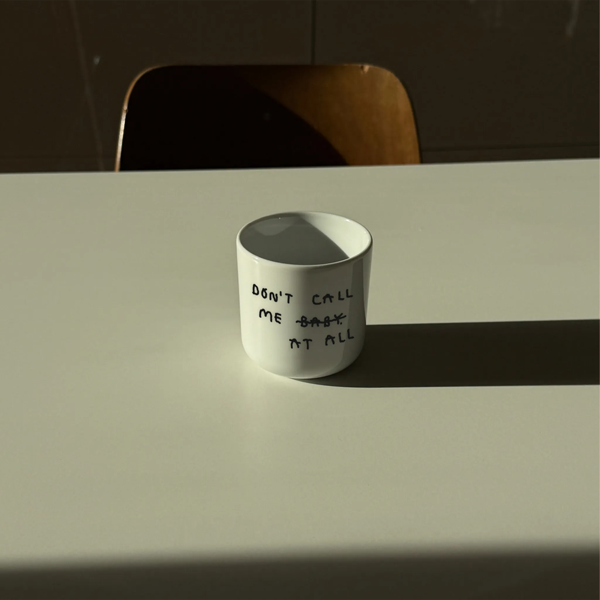 Weißer henkelloser Porzellan-Becher mit schwarzen Text „don't call me at baby" - das Baby ist durchgestrichen und durch "at all" ersetzt. Zu Deutsch (Nenn mich nicht Baby, bzw. Ruf mich nicht an). Der Becher steht auf einem Tisch in angenehmen Licht, ein Holzstuhl ist im Hintergrund. 