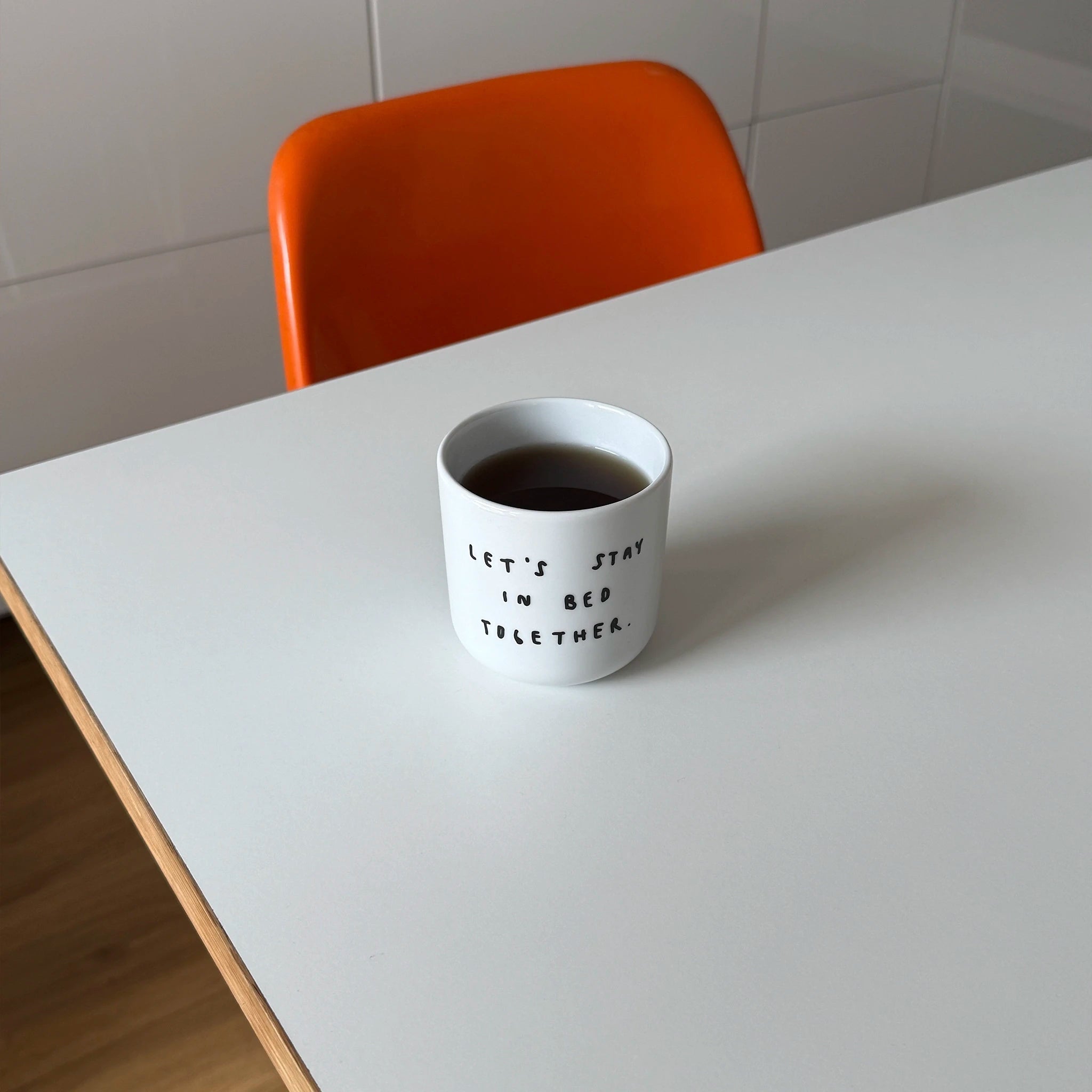 Weißer henkelloser Porzellan-Becher mit einer einfachen schwarzen Illustration, einem handschriftlichen Text der besagt "LET'S STAY IN BED TOGETHER" auf einem weißen Tisch mit orangenen Stuhl.
