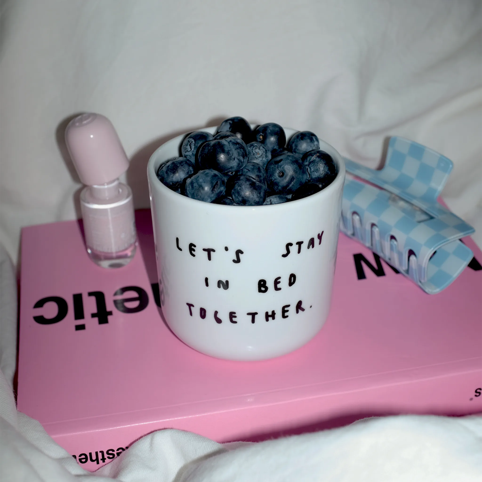 Weißer henkelloser Porzellan-Becher mit einer einfachen schwarzen Illustration, einem handschriftlichen Text der besagt "LET'S STAY IN BED TOGETHER" auf einem pinken Buch. Der Becher ist mit Blaubeeren befüllt, daneben stehen Nagellack und eine Haarklammer.