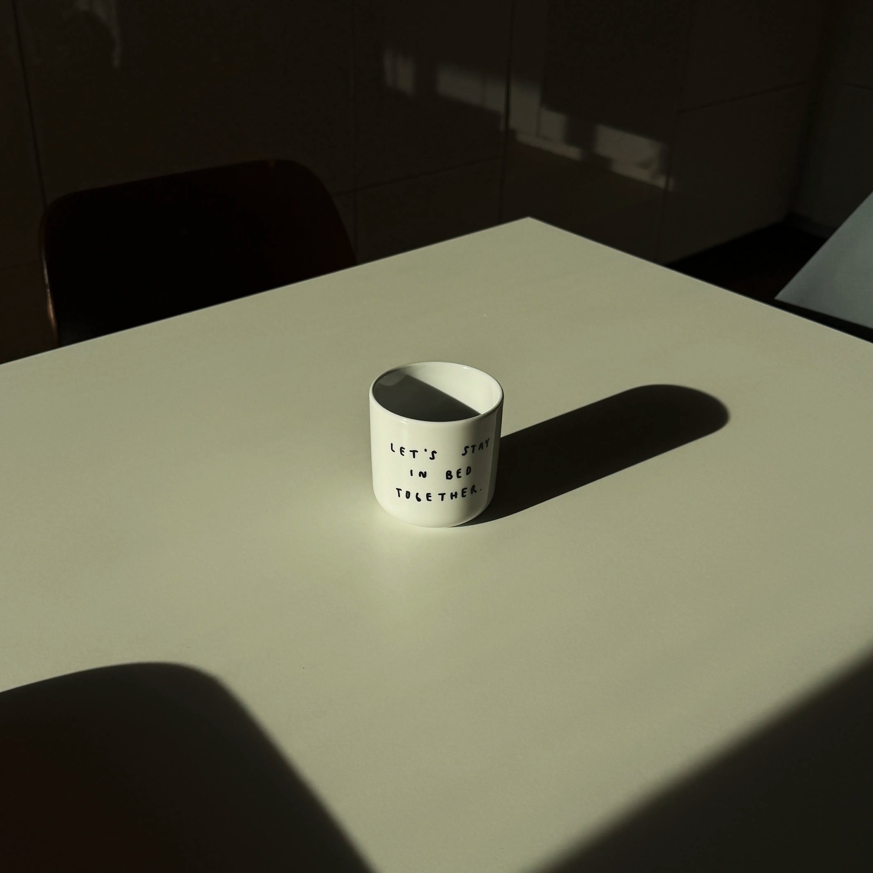 Weißer henkelloser Porzellan-Becher mit einer einfachen schwarzen Illustration, einem handschriftlichen Text der besagt "LET'S STAY IN BED TOGETHER". Der Becher ist nicht befüllt und steht in einem sanften Licht auf einem Küchentisch.