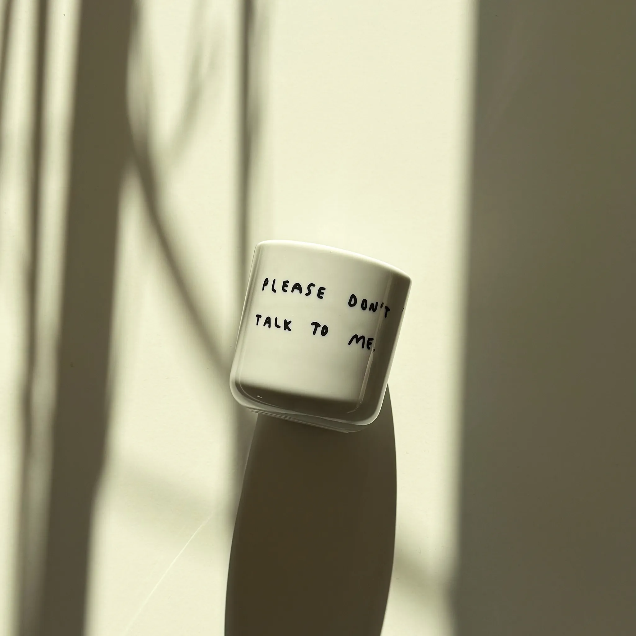 Weißer henkelloser Porzellan-Becher mit einer einfachen schwarzen Illustration, einem handschriftlichen Text der besagt "PLEASE DON'T TALK TO ME" auf weißen Hintergrund liegend. Der Becher liegt in einem milden Licht und wirft einen langen Schatten.