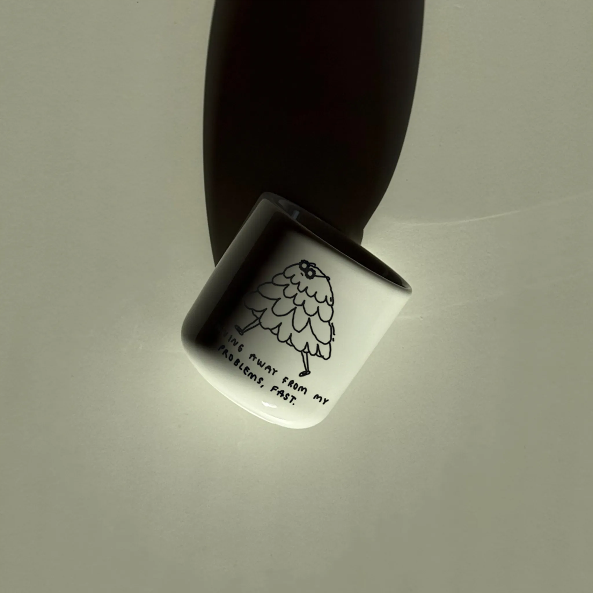 Weißer henkelloser Porzellan-Becher mit einer schwarzen Cartoon-Zeichnung einer flauschigen Figur und dem Text „Running away from my problems, fast“. Der Becher liegt auf einem hellen Tisch im Sonnenlicht und wirft einen langen Schatten.