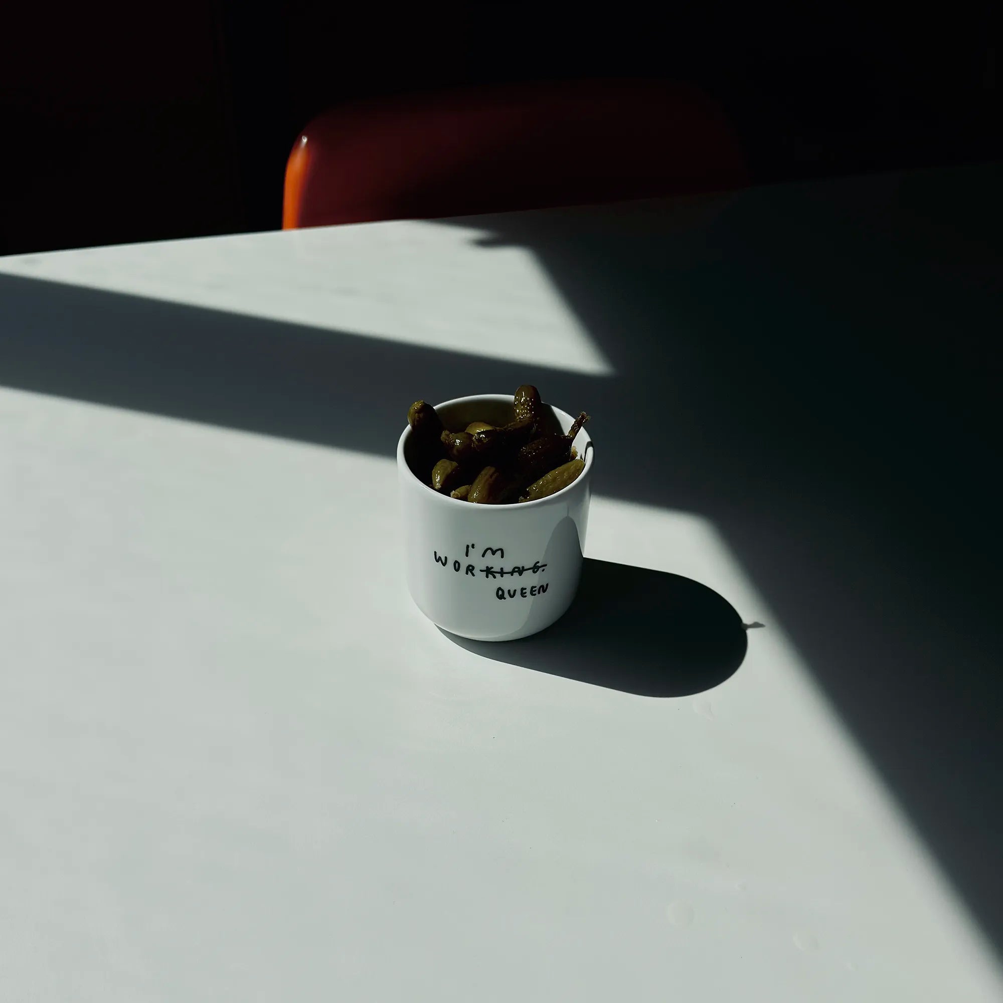 Weißer henkelloser Porzellan-Becher mit einer minimalistischen schwarzen Illustration in der in Handschriftartiger Typografie I'M WORKING steht. Das KING wurde durchgestrichen und unten drunter wurde QUEEN gestrichen. 

Der Becher steht auf einem weißen Tisch und ist mit Gurken befüllt. 