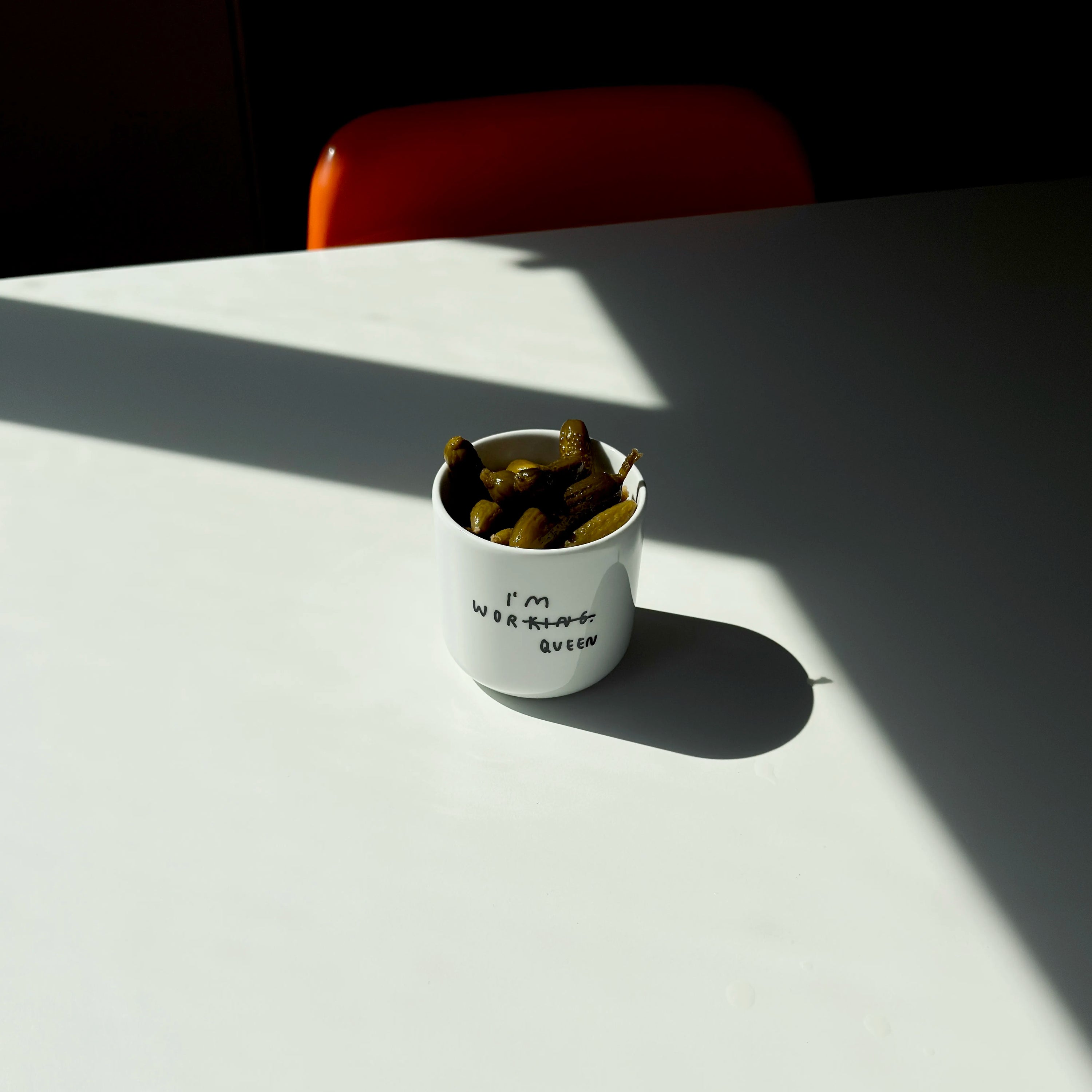 Weißer henkelloser Porzellan-Becher mit einer minimalistischen schwarzen Illustration in der in Handschriftartiger Typografie I'M WORKING steht. Das KING wurde durchgestrichen und unten drunter wurde QUEEN gestrichen. 

Der Becher steht auf einem weißen Tisch und ist mit Gurken befüllt. 
