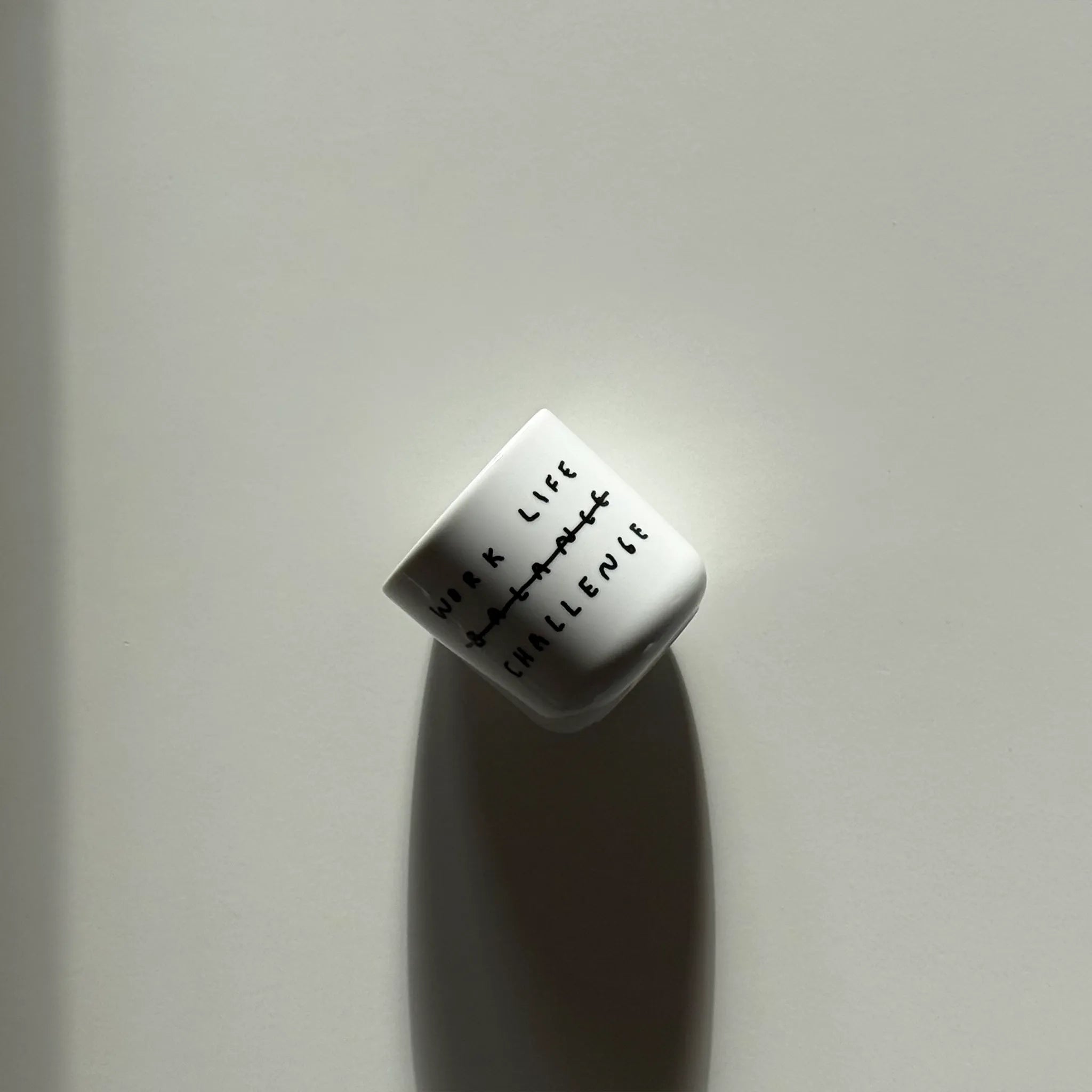 Weißer henkelloser Porzellan-Becher mit einer einfachen schwarzen Illustration, einem handschriftlichen Text der besagt "WORK LIFE CHALLENGE" wobei auch das Wort Balance dabeisteht und humorvoll durchgestrichen wird, dadurch von Challenge ersetzt wird. Der Becher steht in einem Regal zwischen zwei Büchern. 
