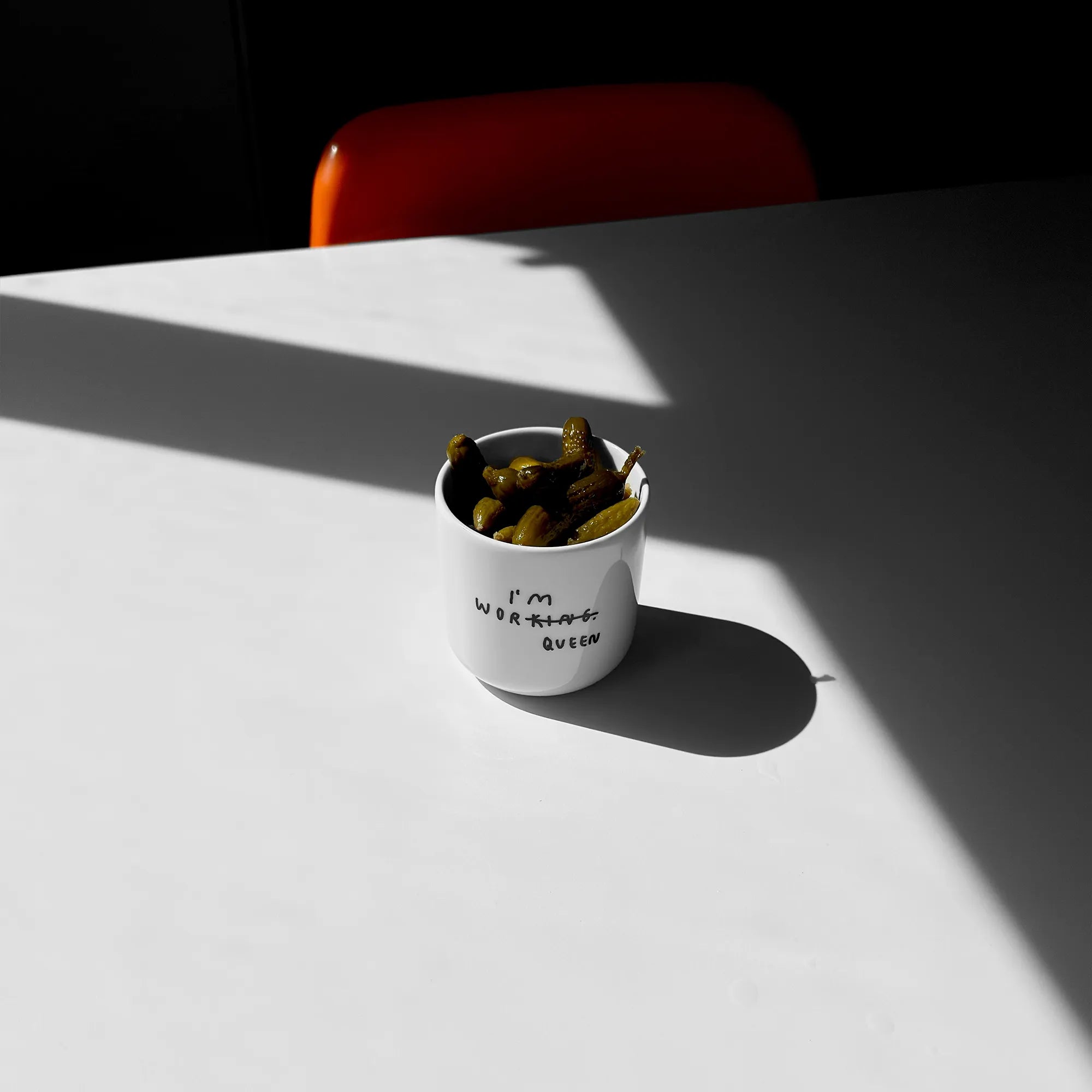 Weißer henkelloser Porzellan-Becher mit einer minimalistischen schwarzen Illustration in der in Handschriftartiger Typografie I'M WORKING steht. Das KING wurde durchgestrichen und unten drunter wurde QUEEN gestrichen. 

Der Becher steht auf einem weißen Tisch und ist mit Gurken befüllt. 
