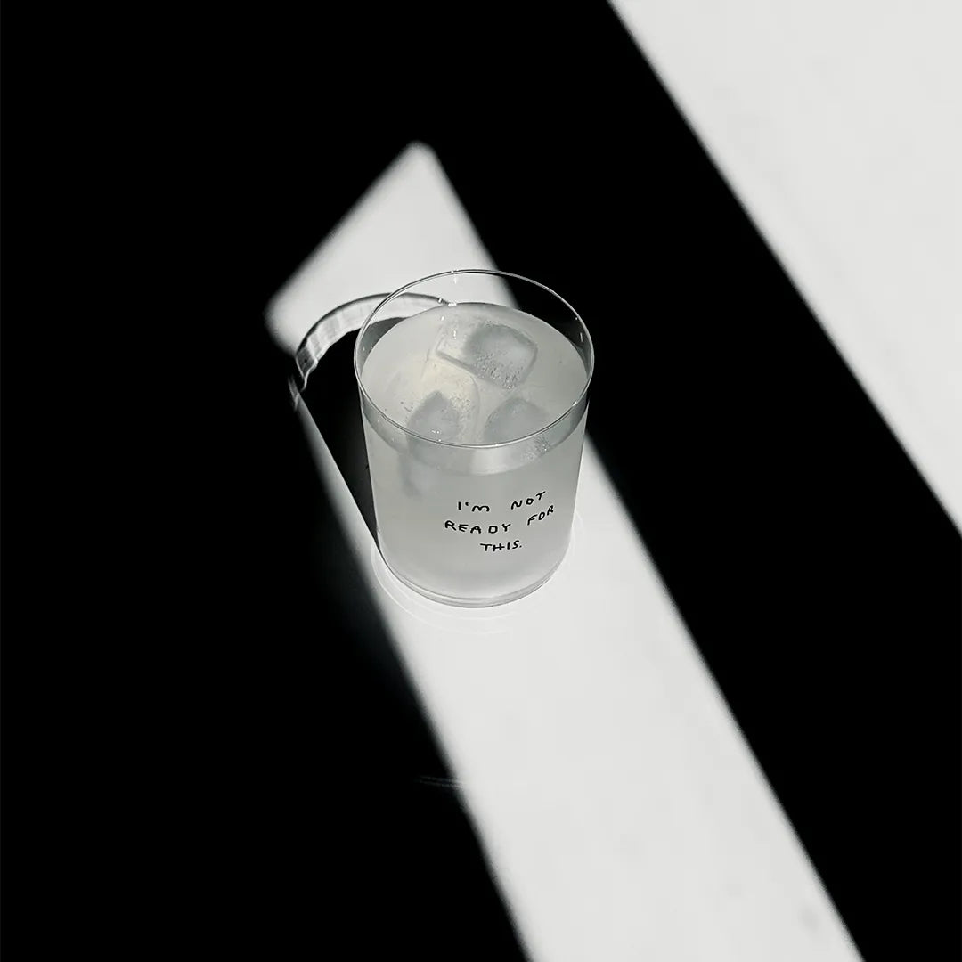 Transparentes Medium Designer Glas mit schwarzem Schriftzug i'm not ready for this". Das Glas hat dünne Wände und Boden. Das Glas steht im Licht auf einem weißen Tisch. Der Rest des Bildes ist dunkel in Schatten gefallen.

Das Glas ist befüllt mit einer klaren Flüssigkeit und einem Eiswürfel.