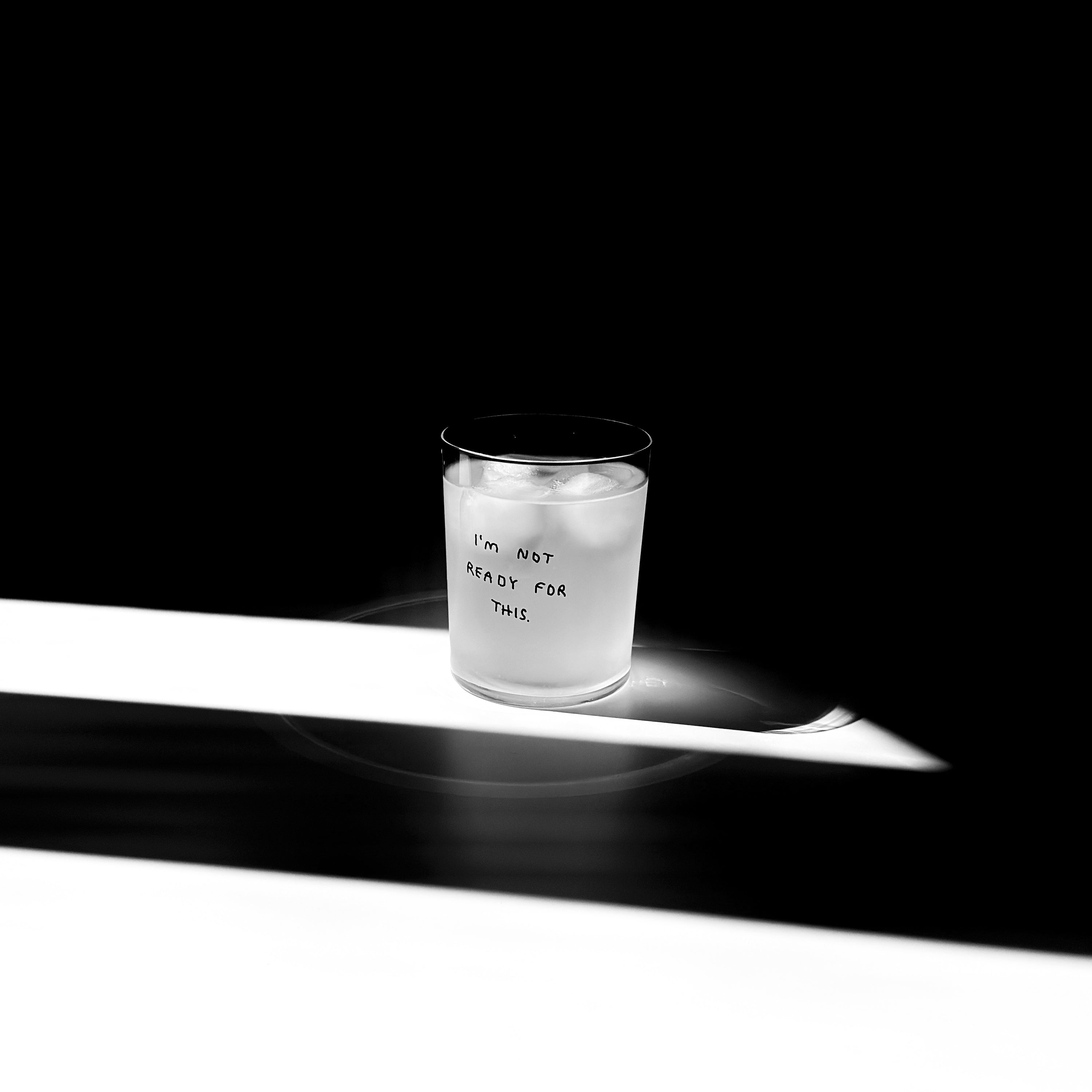 Transparentes Medium Designer Glas mit schwarzem Schriftzug i'm not ready for this". Das Glas hat dünne Wände und Boden. Das Glas steht im Licht auf einem weißen Tisch. Der Rest des Bildes ist dunkel in Schatten gefallen.

Das Glas ist befüllt mit einer klaren Flüssigkeit und einem Eiswürfel.